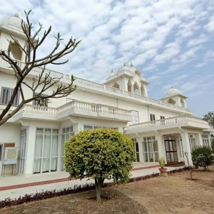 A view of a Savista Retreat haveli white villa with traditional architecture, featuring a large tree in the foreground and a clear blue sky overhead
