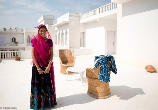 A medium shot of a woman in traditional Rajasthani clothing standing on a white terrace at the Savista Retreat. She is wearing a bright pink headcloth and a long, patterned skirt. Behind her are chairs and a small table.