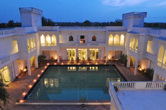 An aerial view of the Savista Retreat at dusk. The central element is a large rectangular pool reflecting lights from the surrounding white building in traditional rajasthani architecture.