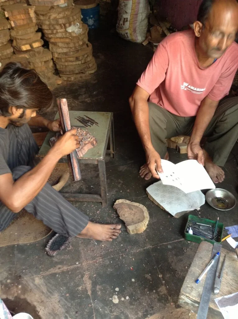 Craftsmen in a rustic workshop; one focused on wood carving, the other checking plans for detailed traditional work