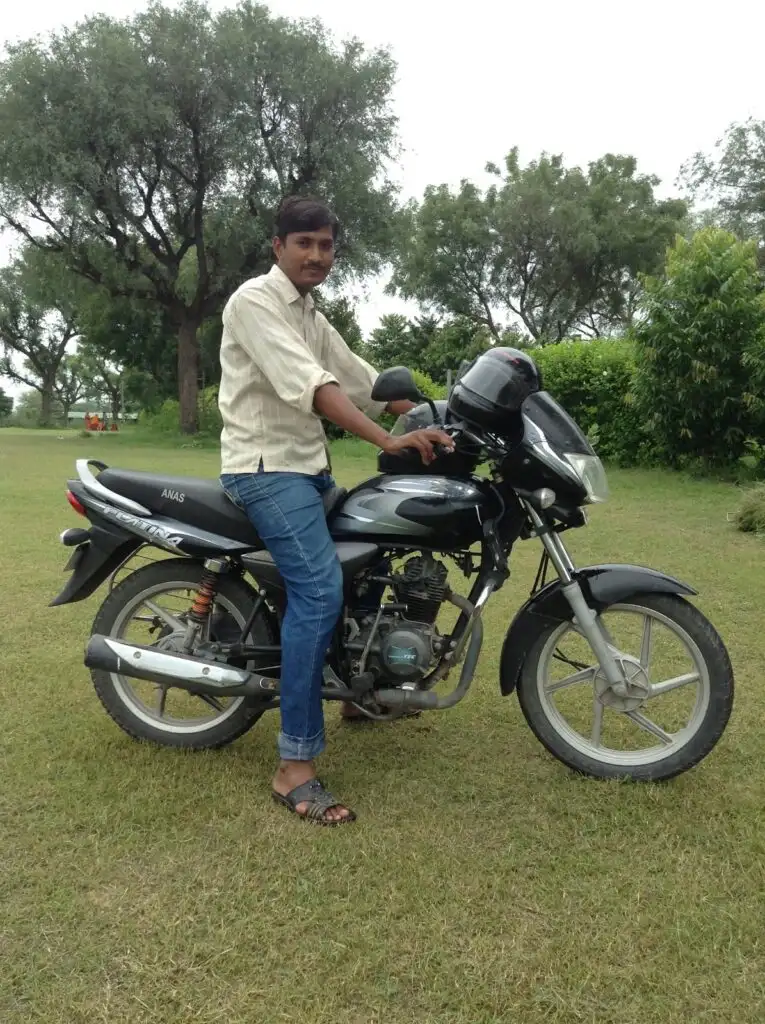 A person wearing a white shirt and sandals, standing on a black motorcycle in a grassy field with trees in the background.