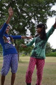two girls joyfully engaging each other with arms wide. They wear pink and blue clothing.