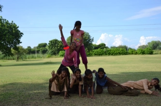 A wide shot shows six young girls outdoors on a bright day, forming a human pyramid on a grassy field at Savista Retreat.