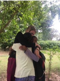 The image shows two women embracing under the shade of a large leafy tree.