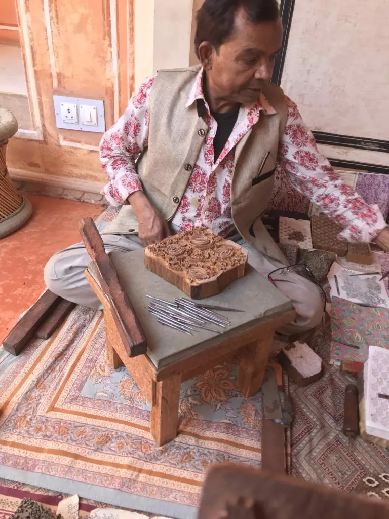 At Savista Retreat, an artisan is shown seated on the floor working on a wood block, with tools and other wood blocks on a low stool, demonstrating traditional Indian textile printing.