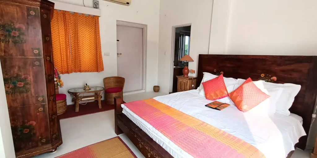 Another view of the bedroom with a wooden wardrobe and a wooden bed with orange and pink bedding, and wicker chairs next to the window.