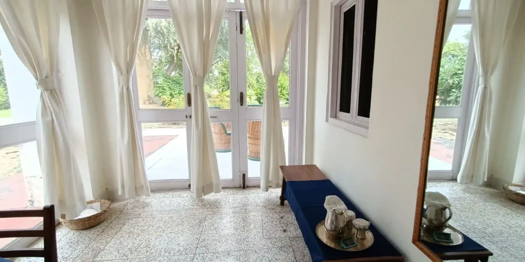 A room corner with a wooden bench and a plant beside a window with light curtains.