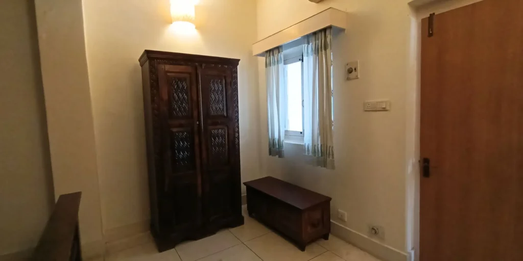 A room with a wooden wardrobe, a bench, and a window with light-colored curtains.