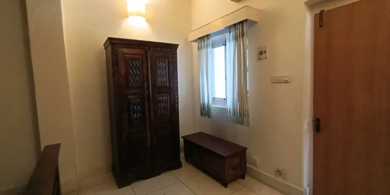 A room with a wooden wardrobe, a bench, and a window with light-colored curtains.