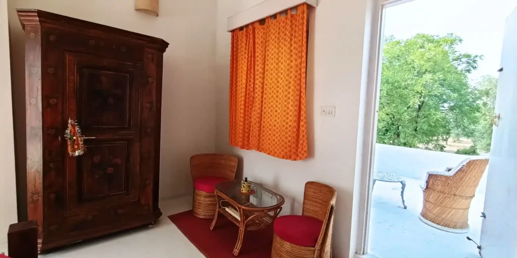 A cozy corner of the room with a wooden wardrobe, wicker chairs, a glass coffee table, and a window with orange curtains.