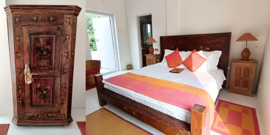 A close-up of a floral-painted wardrobe with a small wooden bedside table, wooden bed, and a clear view of the outside from the window.