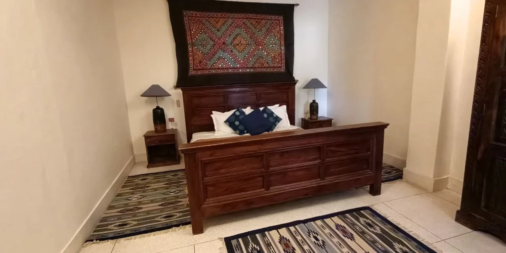 A bedroom with a wooden bed, blue cushions, and an ethnic wall hanging above the headboard.