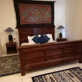 A bedroom with a wooden bed, blue cushions, and an ethnic wall hanging above the headboard.