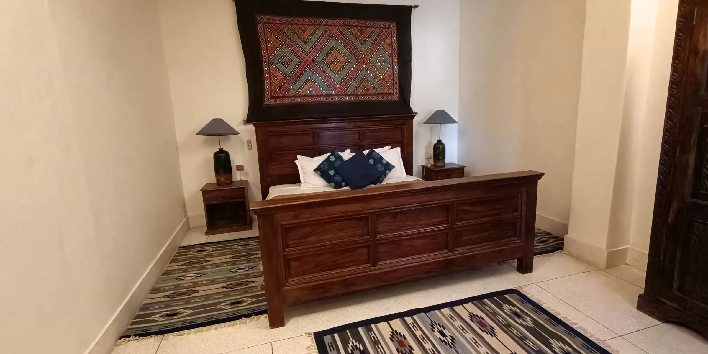 A bedroom with a wooden bed, blue cushions, and an ethnic wall hanging above the headboard.