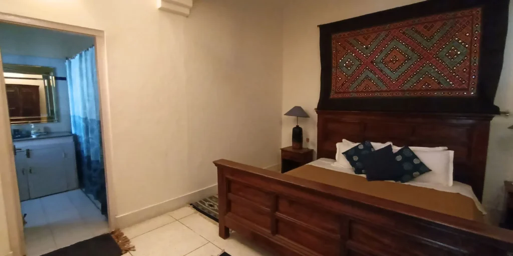 A bedroom featuring a wooden bed with ethnic bedding, a decorative wall hanging, and a bathroom doorway visible.