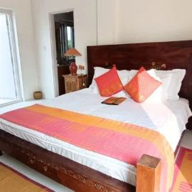 A well-lit bedroom featuring a wooden bed with vibrant pillows, a small wooden nightstand, and a view of the outdoors through an open door.