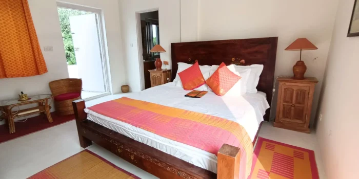 A well-lit bedroom featuring a wooden bed with vibrant pillows, a small wooden nightstand, and a view of the outdoors through an open door.