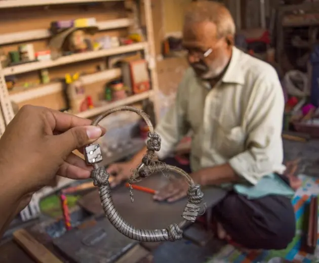 A hand holding a shaped bangle in a workshop, with a man working in the background.