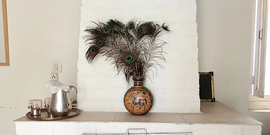 A decorative vase with peacock feathers on a mantelpiece, with a silver pitcher and cups placed on a tray, and a framed artwork on the wall next to the window.