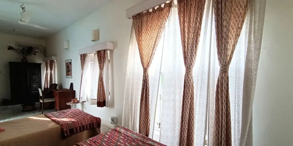 A well-lit bedroom with two single beds, decorated with patterned bedspreads and pillows, and window curtains letting in natural light.