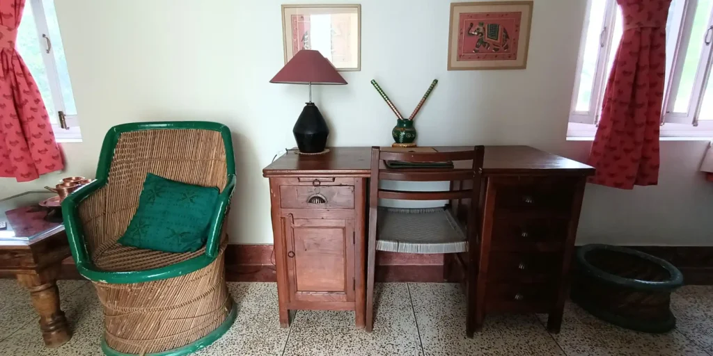 A room with a wicker chair, wooden desk, and red curtains by the window.