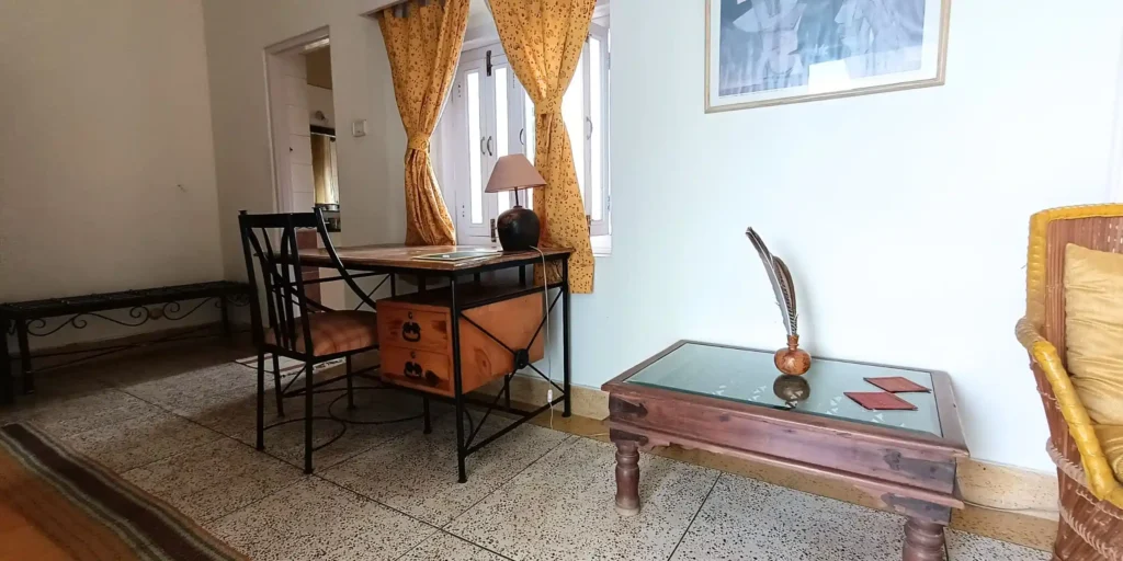 A room with a wooden desk, chair, and a cozy seating area featuring a rattan chair and decorative items on the table.