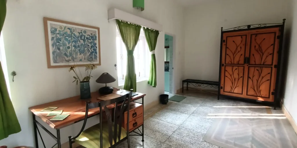 A room featuring green curtains, a wooden desk with a lamp, and a wardrobe in the corner.