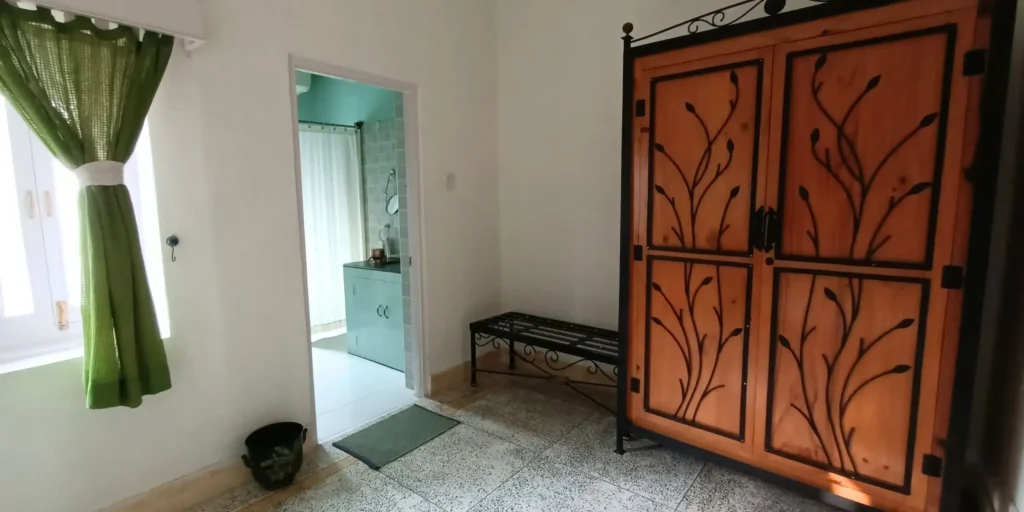 A room with a wooden wardrobe, a bench, green curtains, and a window leading to a bathroom.