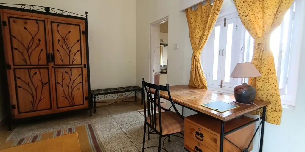 A room with a wooden wardrobe, a study desk with a lamp, and yellow curtains on the windows.