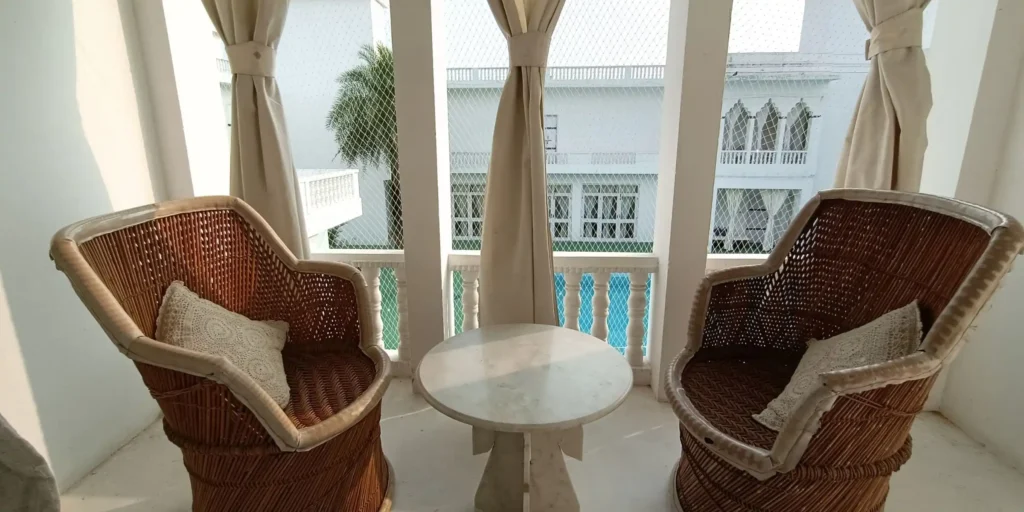 A cozy balcony area with two wicker chairs and a round marble table, with sheer curtains and a view of a pool and white building in the background.