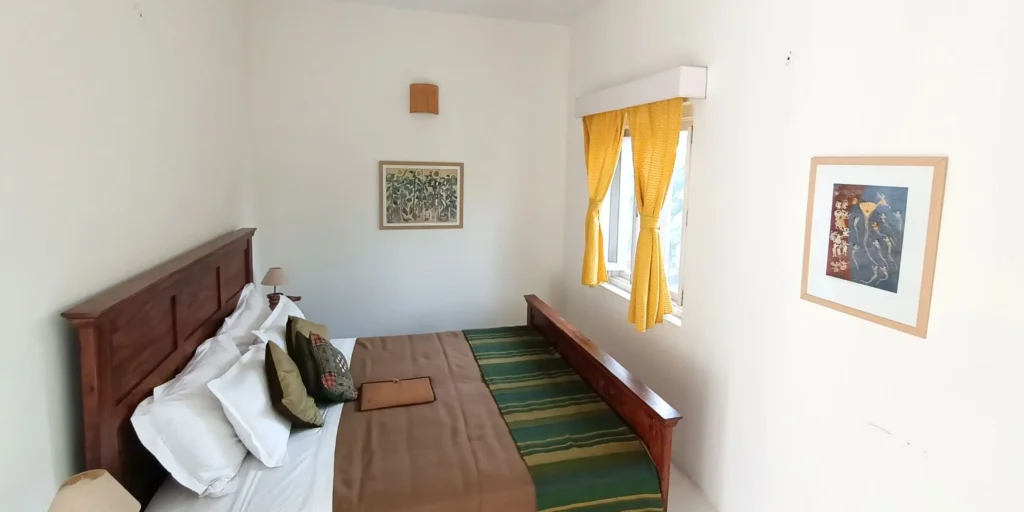 A cozy bedroom featuring a wooden bed with a green and yellow bedspread, a peacock feather vase on the nightstand, and bright yellow curtains framing the window.