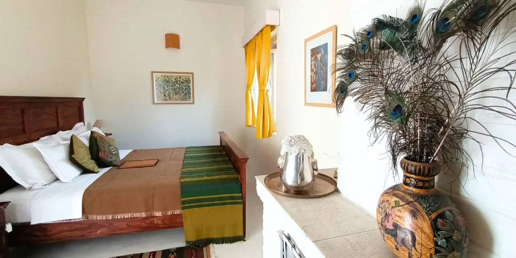 A bedroom with a wooden bed, green and yellow bedspread, a peacock feather vase on a table, and framed artwork on the walls. Yellow curtains complement the natural light coming in.