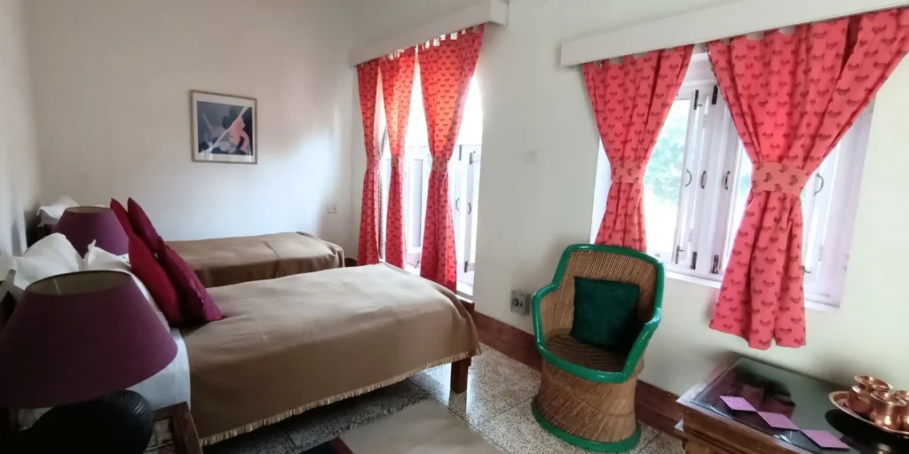 A room featuring twin beds with brown blankets, red curtains, and green wicker chairs by the window.