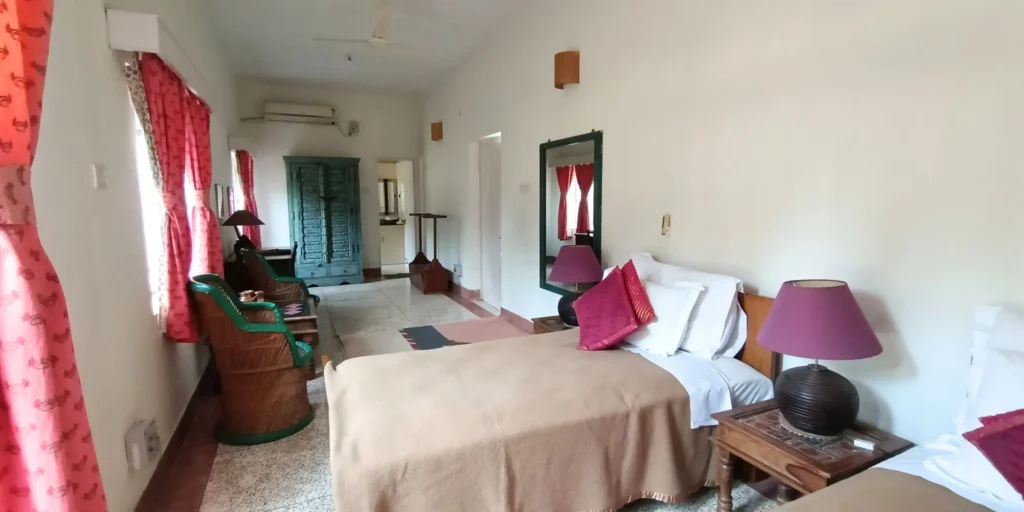 A room with twin beds, red curtains, green wicker chairs, and a blue wooden wardrobe.