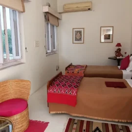 A well-lit bedroom with two single beds, vibrant patterned bed covers, and a wicker chair beside a small table.