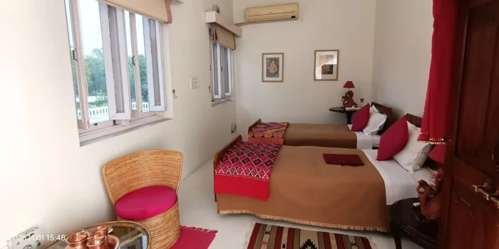 A well-lit bedroom with two single beds, vibrant patterned bed covers, and a wicker chair beside a small table.