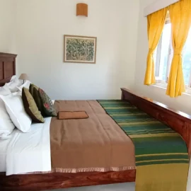 A well-decorated bedroom with a wooden bed, green and yellow bedspread, white pillows, and artwork on the wall. The window has yellow curtains with sunlight streaming in.