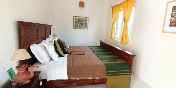 A well-decorated bedroom with a wooden bed, green and yellow bedspread, white pillows, and artwork on the wall. The window has yellow curtains with sunlight streaming in.