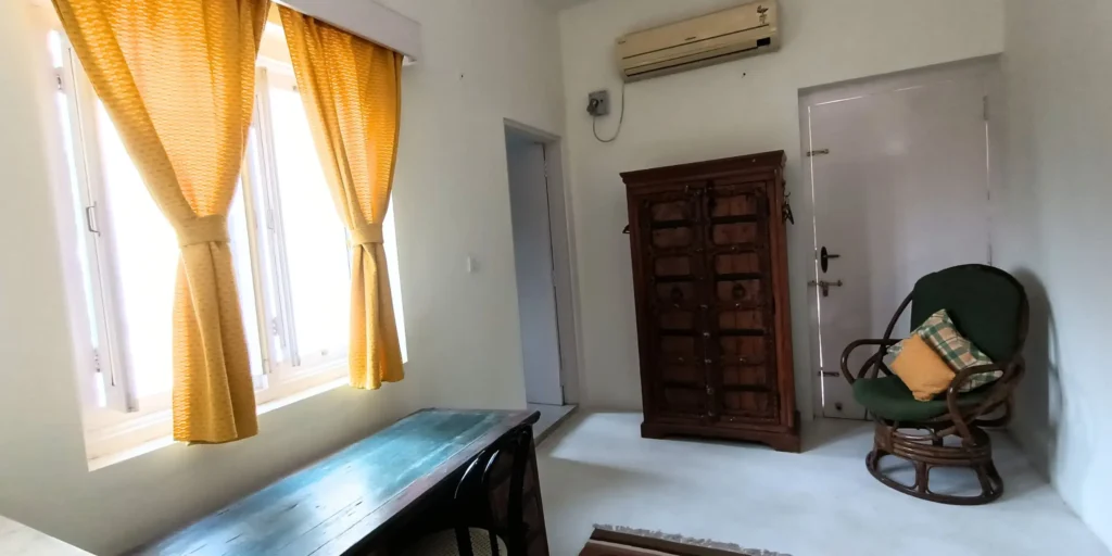 A bedroom with a wooden wardrobe, green rocking chair with colorful pillows, a small desk, and yellow curtains with a bright light coming through the windows.