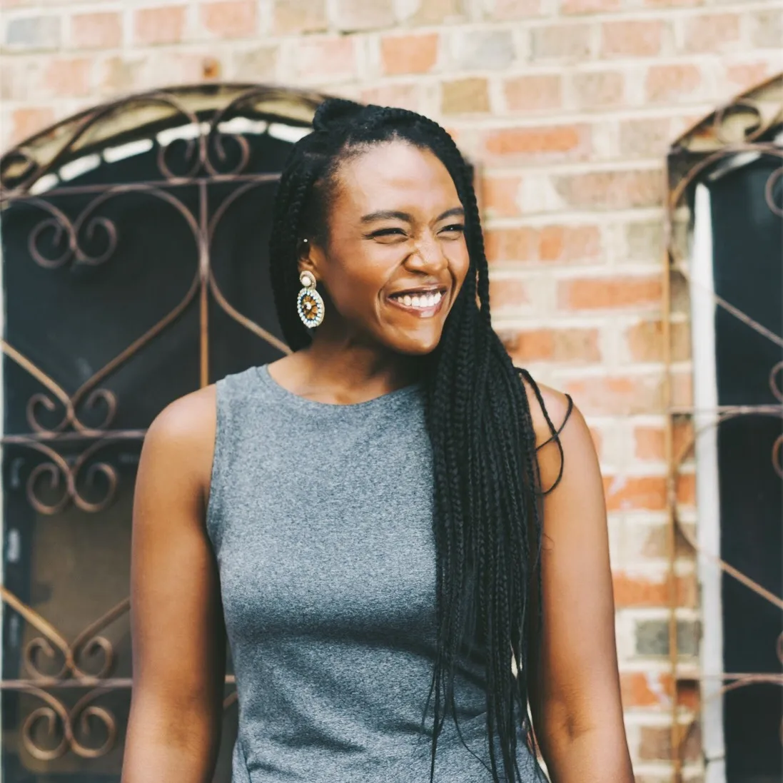 A woman with long braided hair, wearing a grey sleeveless top and large statement earrings, smiling and standing