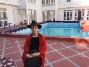 A guest at Savista Retreat sitting by a poolside, wearing a red shawl and black hat, with a white building and clear water in the background.