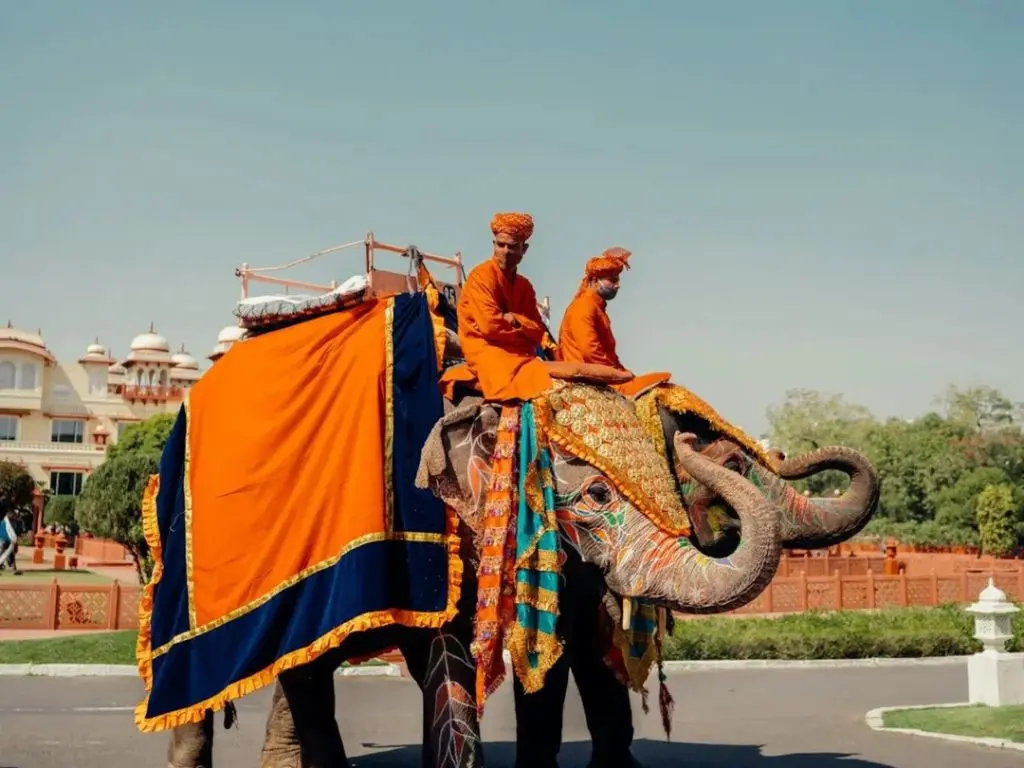 Two riders dressed in orange traditional attire on an adorned elephant in front of a palace.