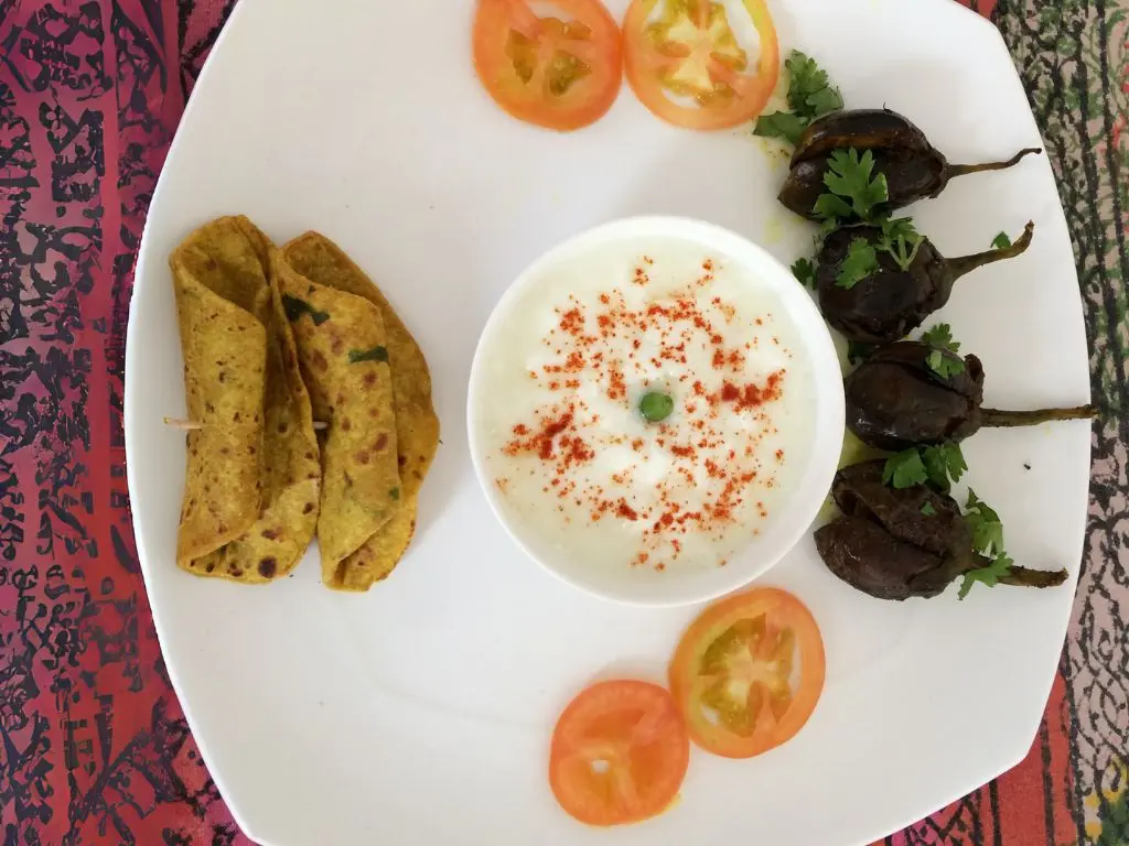 Plate of traditional Indian food featuring stuffed eggplants, yogurt with seasoning, tomato slices, and paratha.