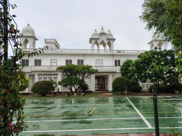 White multi-story building featuring Rajasthani architectural chhatris and a green outdoor sports court in the foreground.