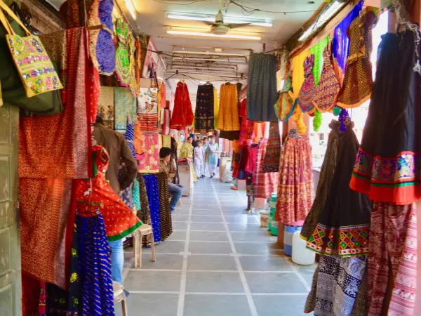 A covered walkway in a vibrant Jaipur bazaar lined with brightly colored traditional Rajasthani textiles and skirts hanging from the shopfronts.