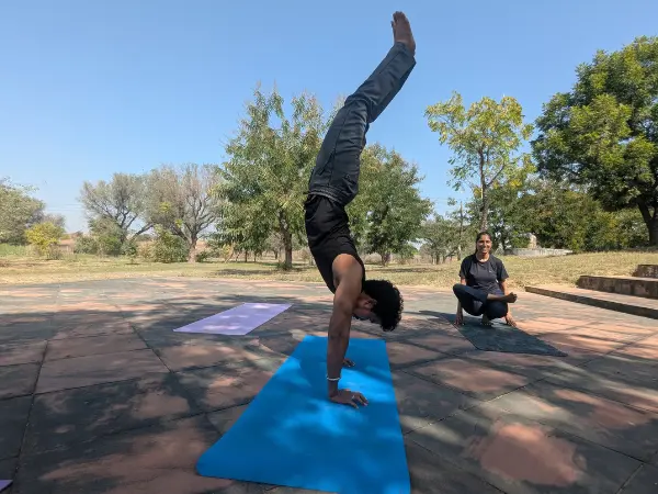 A man performing a full handstand on a blue mat outdoors while a woman sits in a yoga pose on a mat behind him.