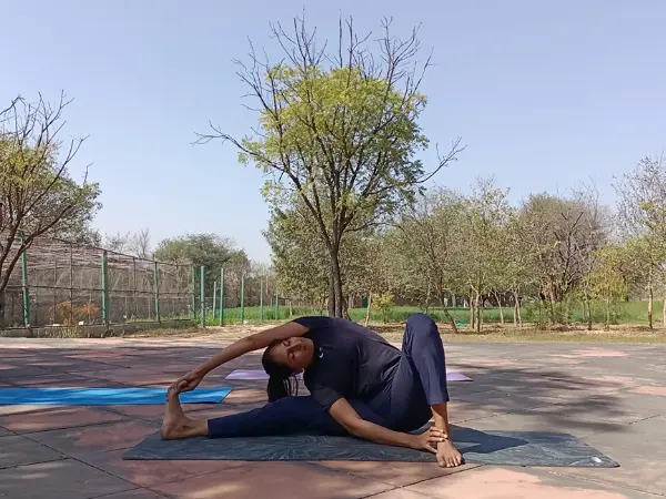 A woman in dark athletic wear performing a seated side stretch yoga pose outdoors on a yoga mat.