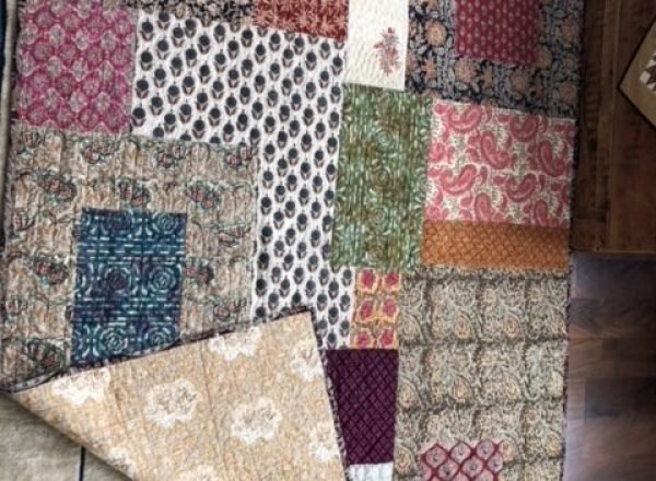 An artisan patchwork quilt featuring a variety of floral and geometric patterns is partially folded back to reveal a tan-colored patterned lining.