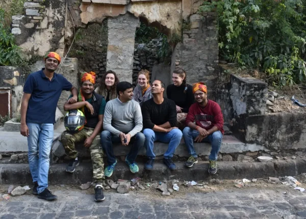 A group of eight travelers pose together, wearing orange turbans, in front of a historical stone structure.