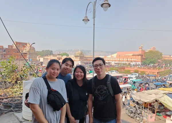 A group of four tourists stands in front of a busy street in Jaipur, with the beautiful backdrop of traditional buildings and traffic.